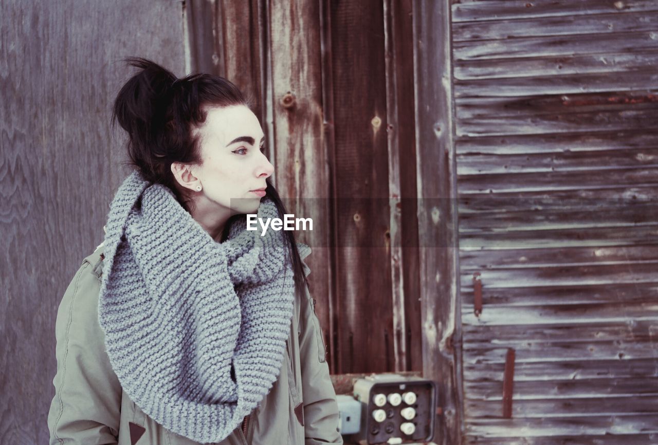 Woman looking away while standing against wooden wall during winter