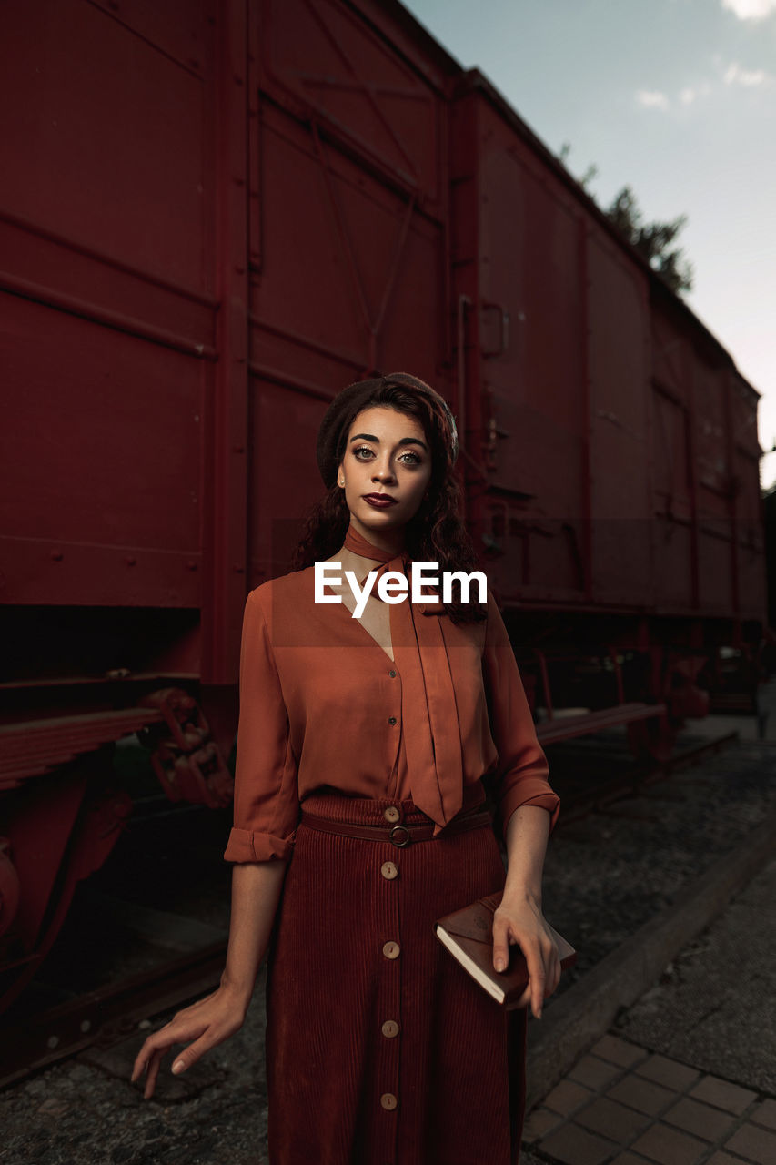 Dark haired female totally wearing terracotta clothes in vintage style standing near terracotta car train and holding open book in hands having dreaming look