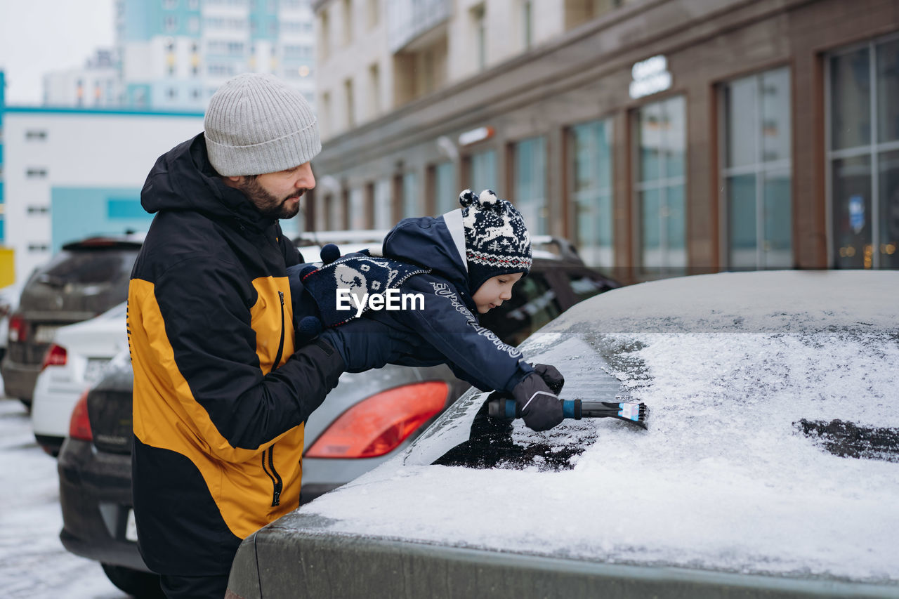 Boy wearing scandinavian knit hat helping to brush snow from a car