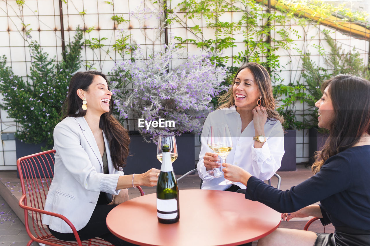 Cheerful adult girlfriends in smart casual clothes sitting at restaurant table and clinking glasses of white wine during meeting on terrace