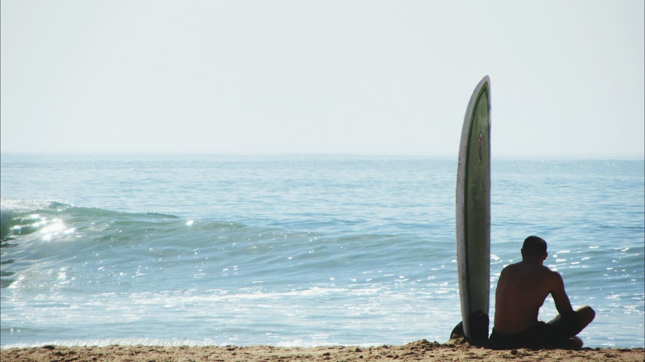 Rear view of man with surfboard sitting on beach against clear sky