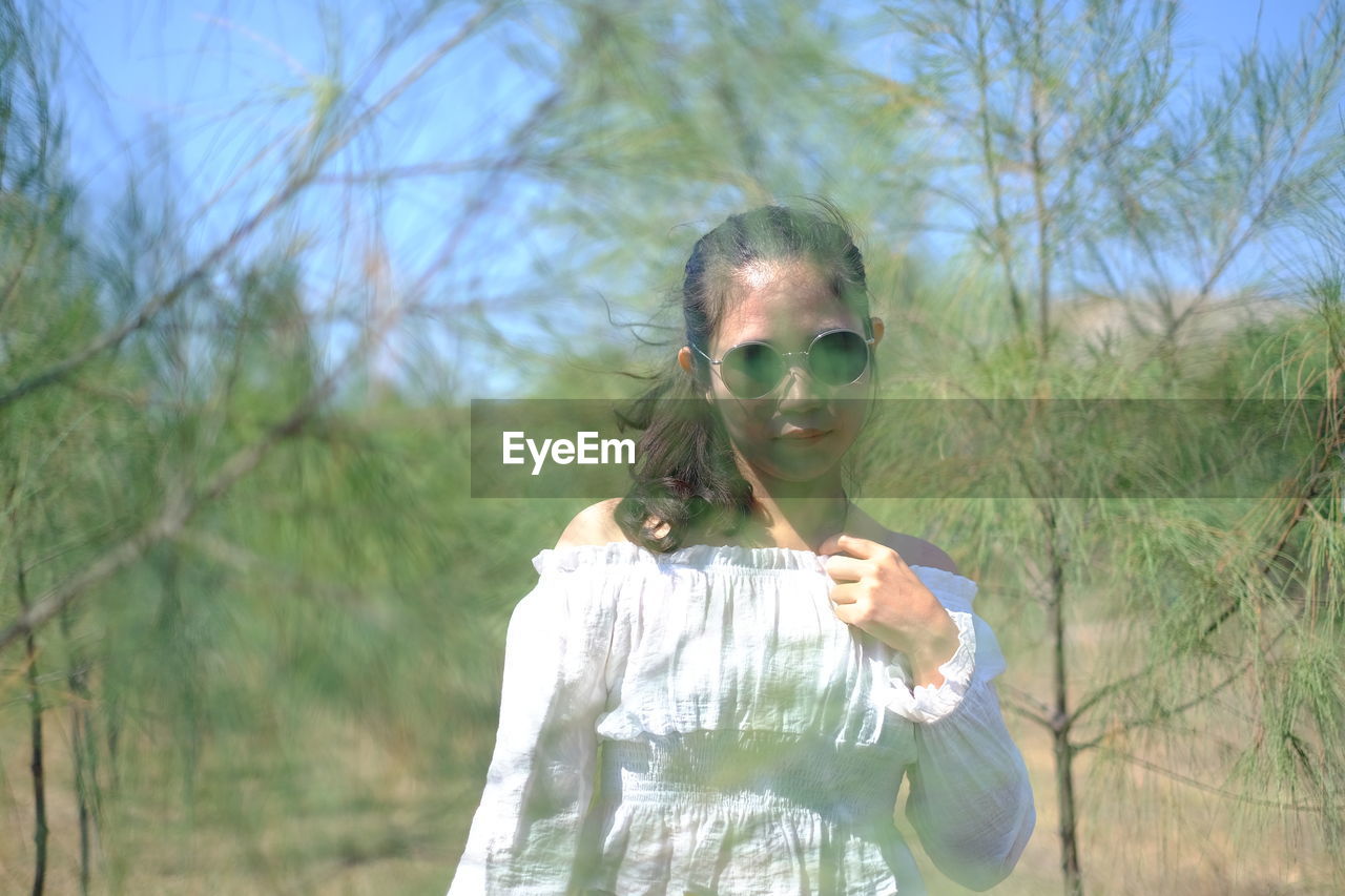 one person, plant, nature, tree, adult, sunglasses, glasses, fashion, portrait, women, young adult, waist up, standing, day, front view, land, spring, leisure activity, sunlight, outdoors, lifestyles, sky, casual clothing, clothing, holding, focus on foreground, female, hairstyle, looking at camera, field, person