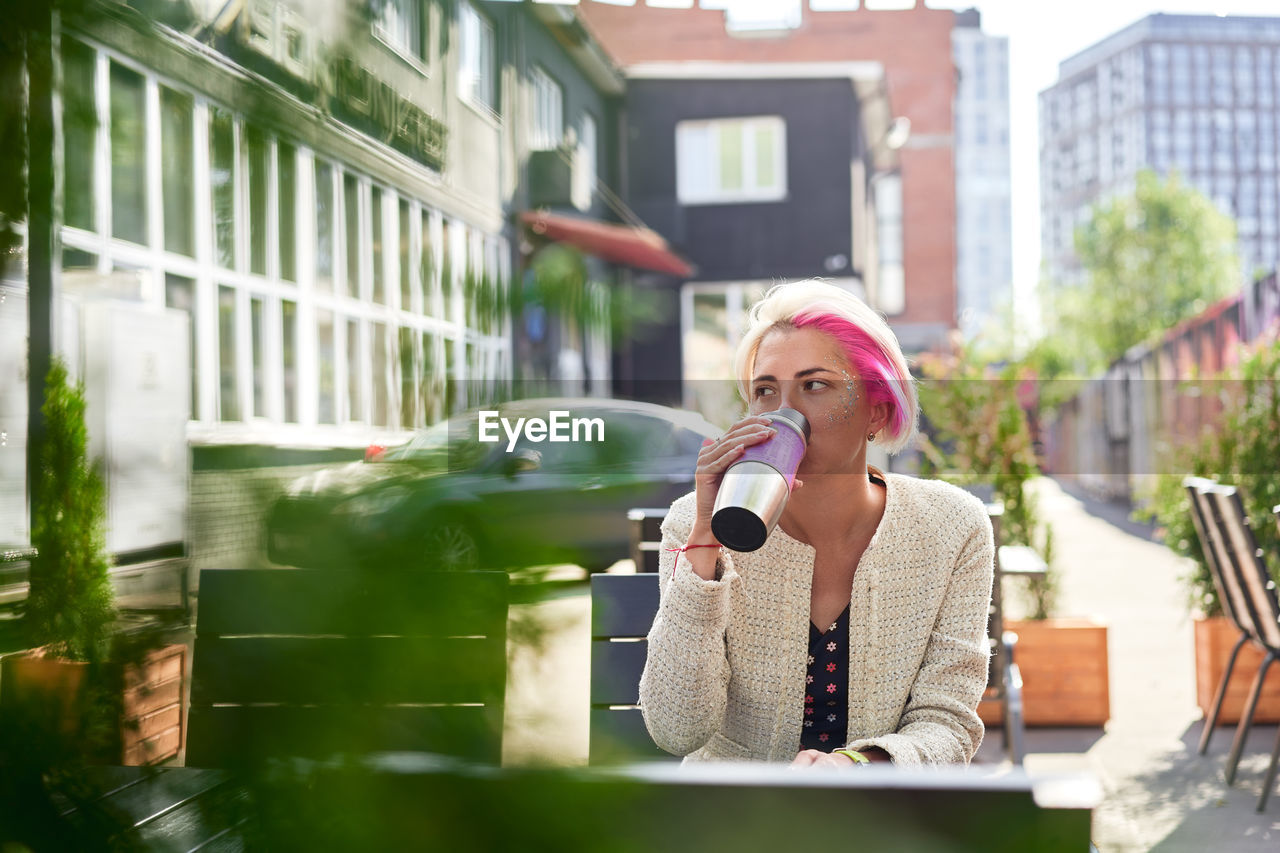 Low angle of pensive alternative female with dyed hair drinking hot beverage from eco friendly cup while sitting at table in street cafe and looking away
