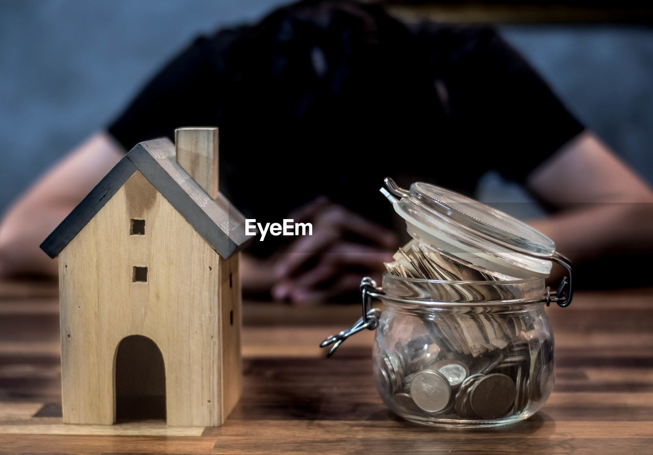 Close-up of model home by savings in jar on table with man in background