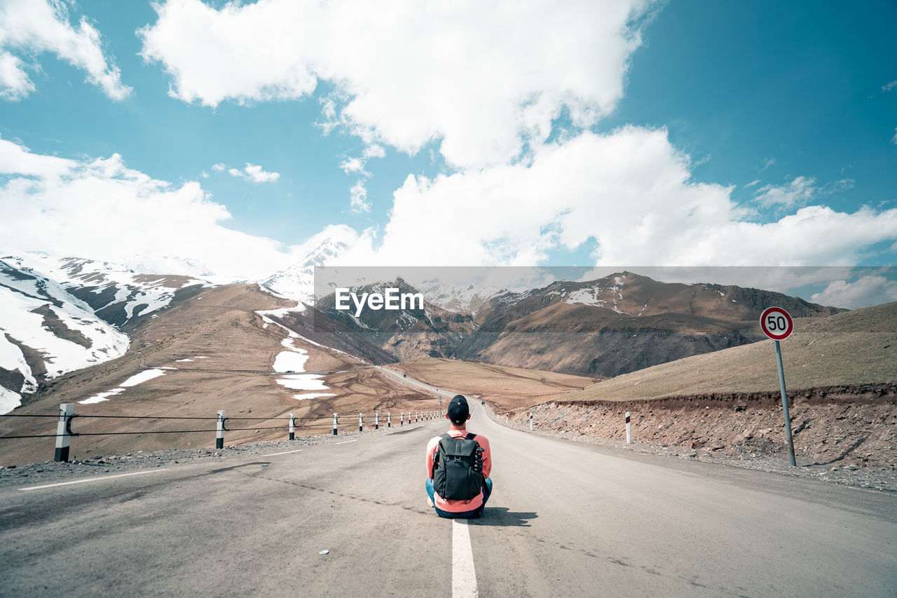 REAR VIEW OF PERSON STANDING ON ROAD AGAINST MOUNTAINS