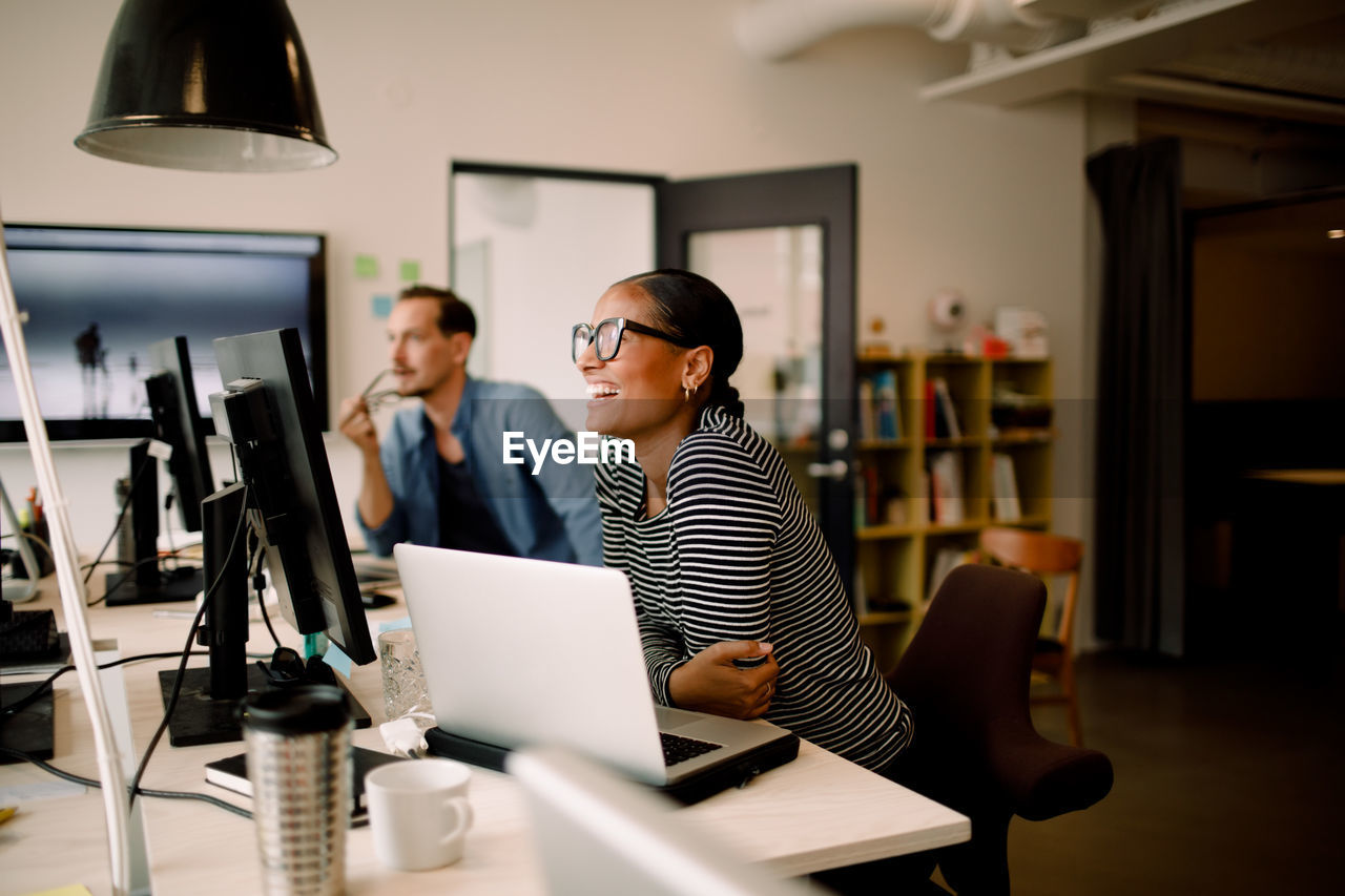 Smiling female colleague sitting by coworker at desk in workplace