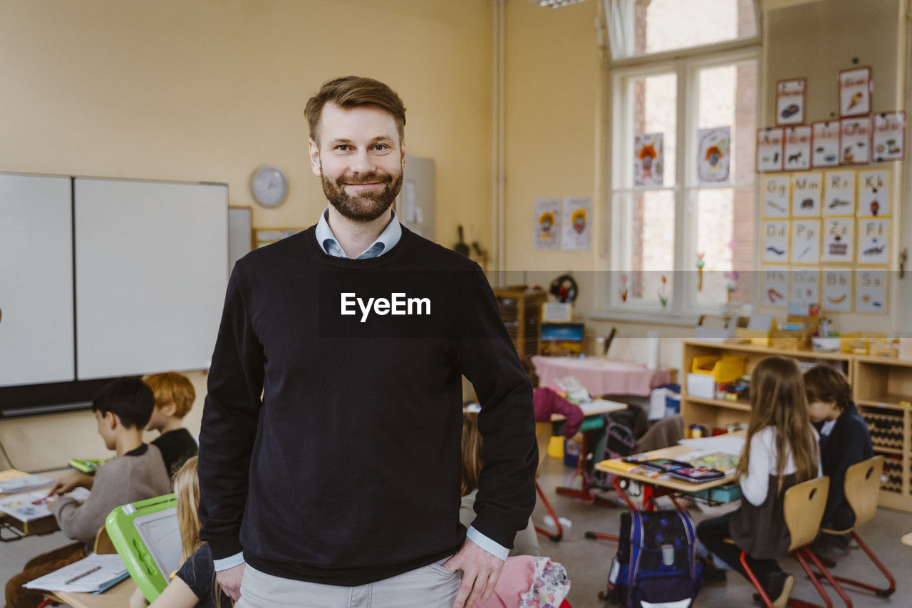 Portrait of smiling bearded teacher standing in front of students at classroom