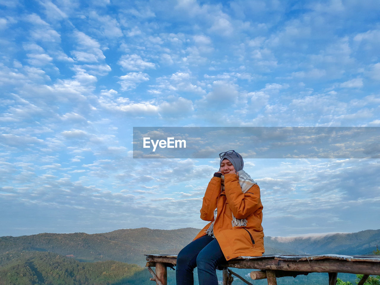 Young woman in warm clothing sitting on observation point against cloudy sky