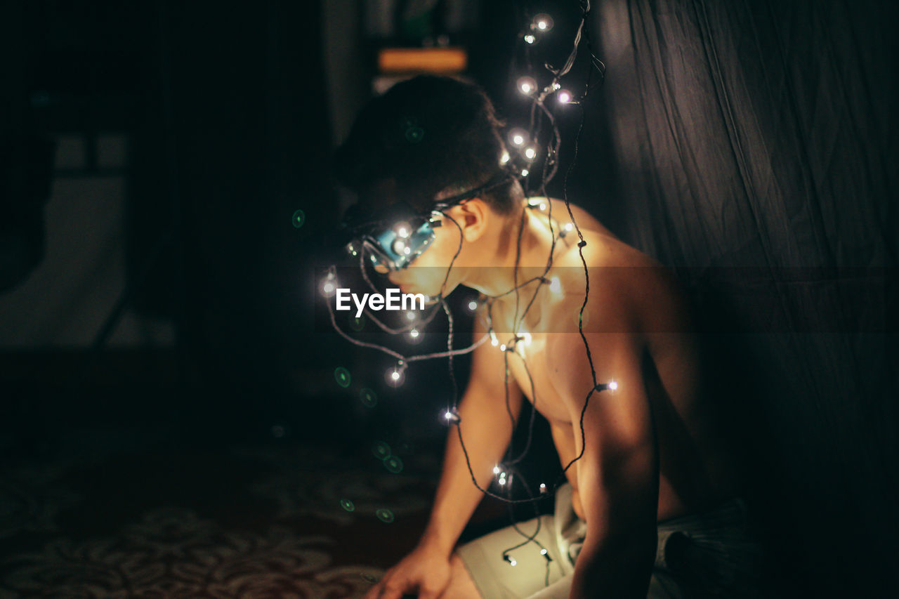 Illuminated string lights on side view of shirtless man sitting at home