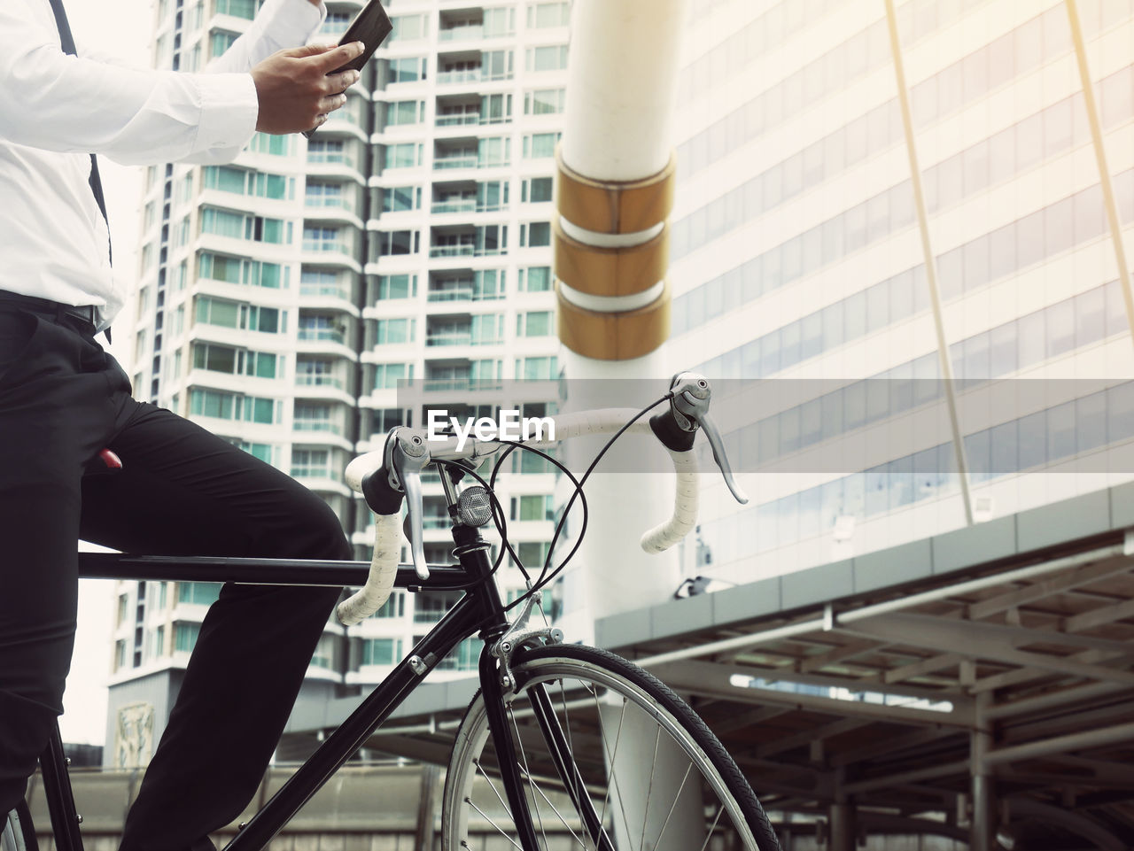 Midsection of businessman with bicycle using smart phone against building in city