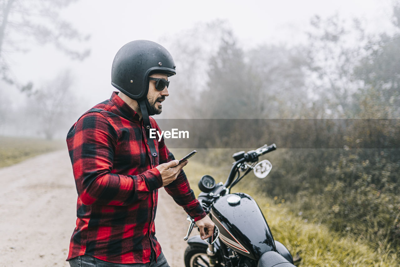 Male biker talking on speaker of smart phone while standing by motorcycle in foggy weather