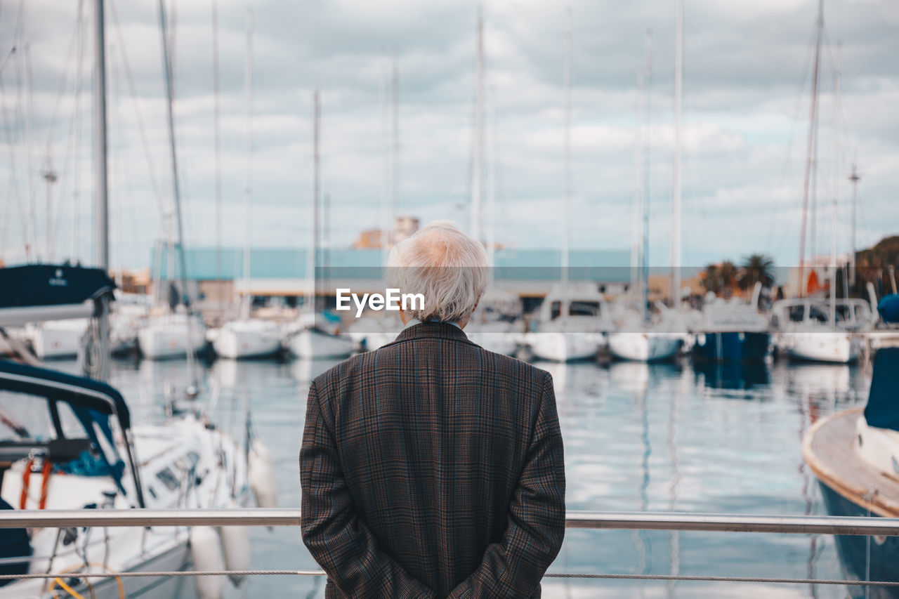 Rear view of senior man with hands behind back standing by railing at harbor