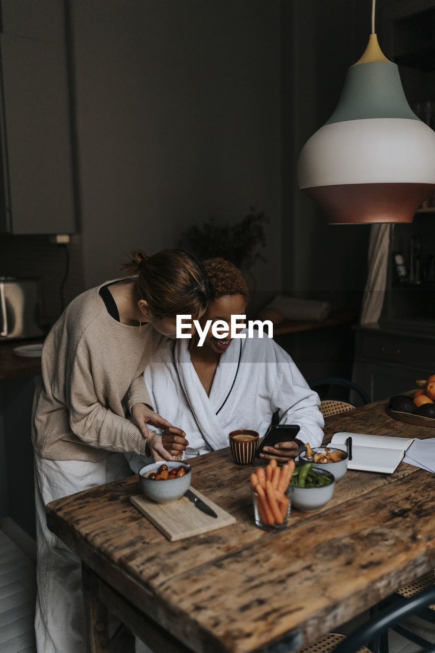 Non-binary couple sharing smart phone while having breakfast at home