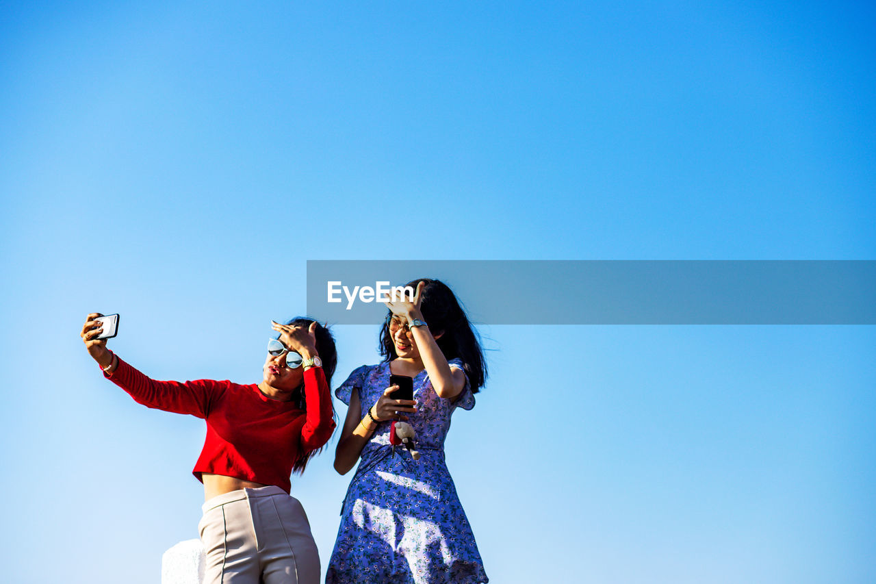 Low angle view of female friends taking selfie while standing against clear blue sky