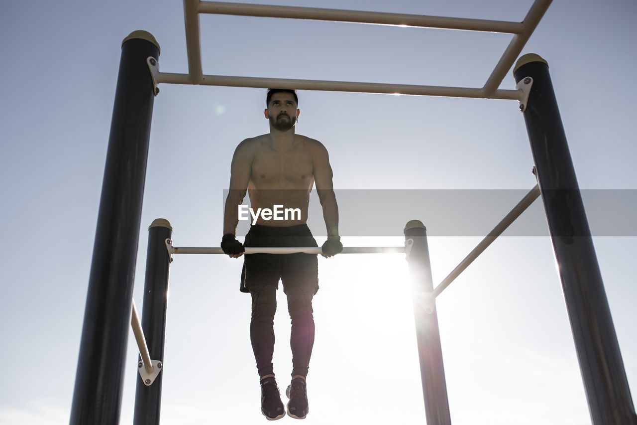 Shirtless male athlete exercising on horizontal bar in park against clear sky