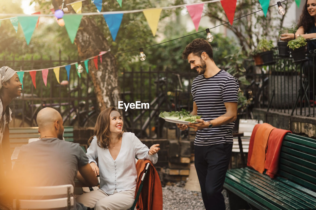 Smiling young woman talking with male friend carrying food during garden party