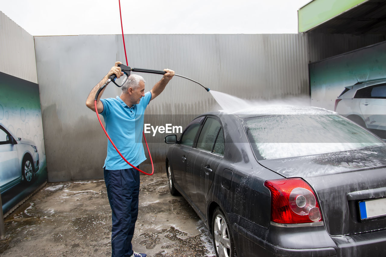 Mature man cleans automobile with covered with foam shampoo chemical detergent carwash self service