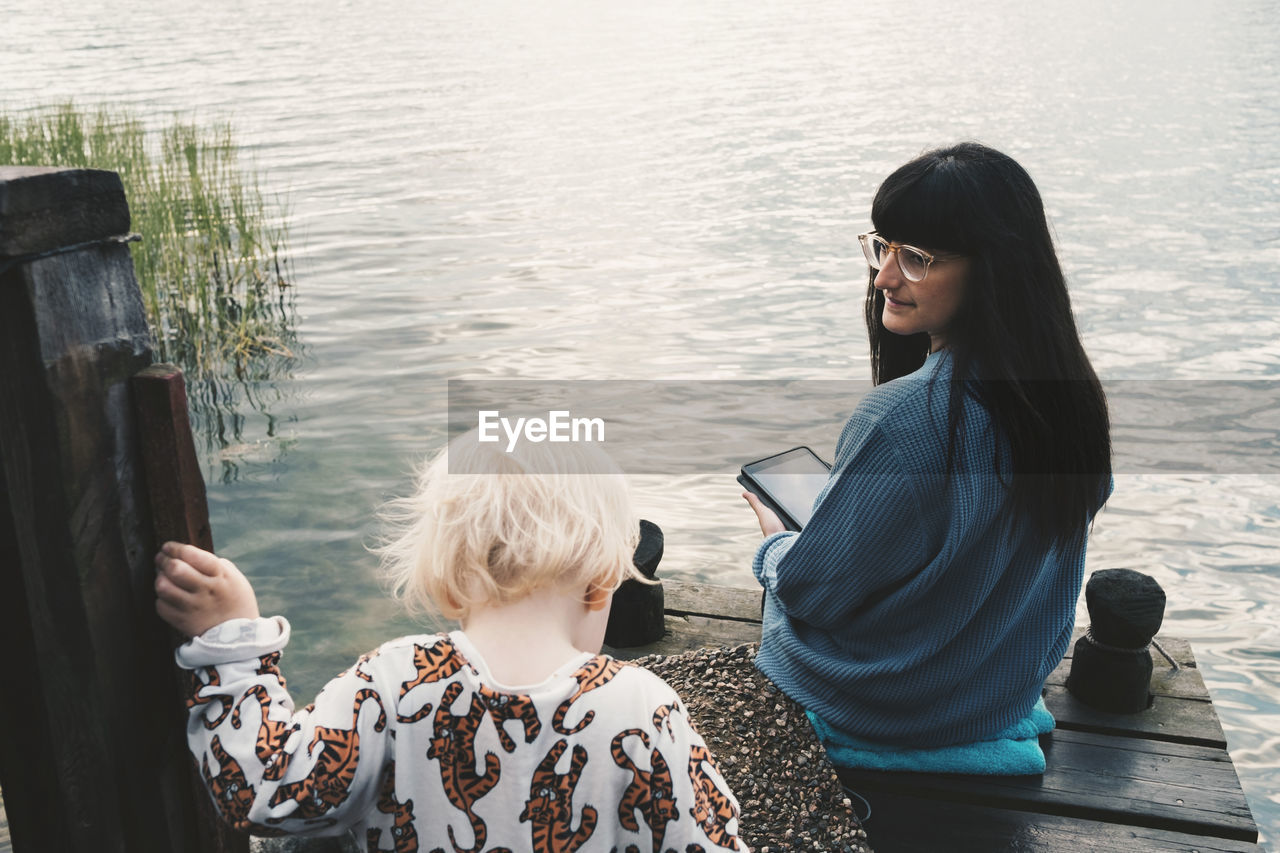 Mother looking at daughter while holding digital tablet and sitting on jetty over lake