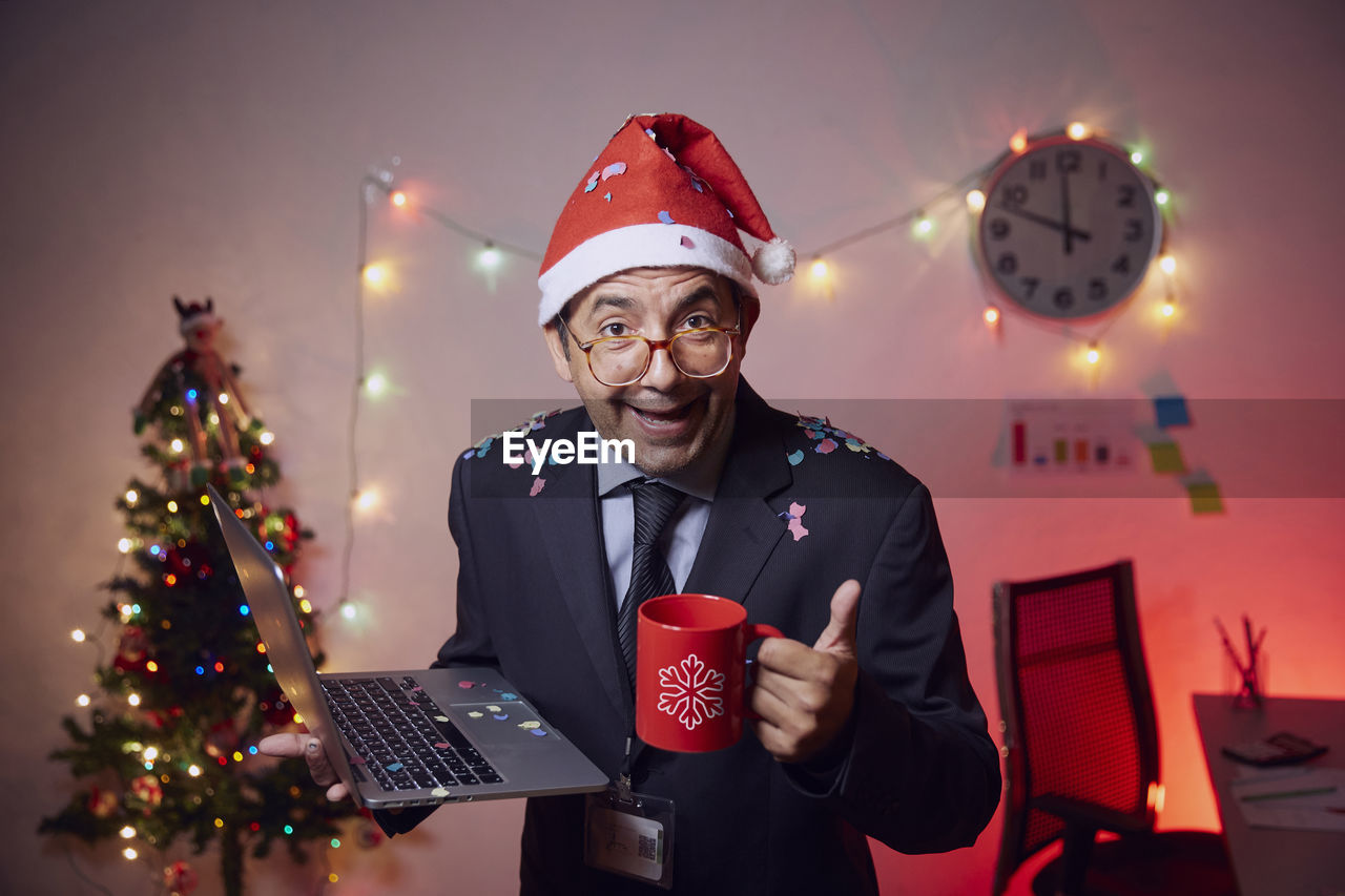 Cheerful businessman wearing santa hat holding laptop and coffee cup against illuminated christmas tree and lights at home