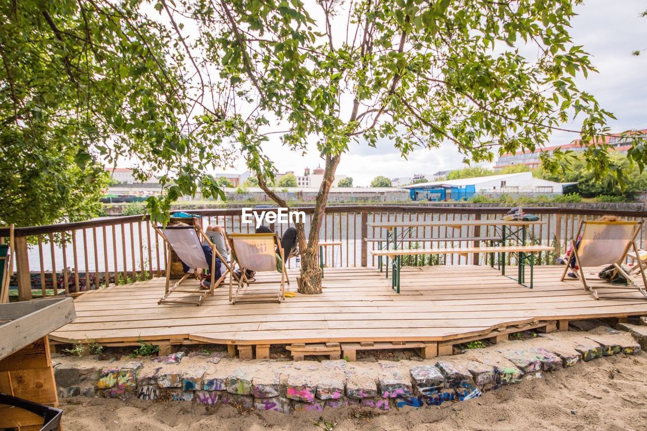 Friends resting on chairs on promenade by trees