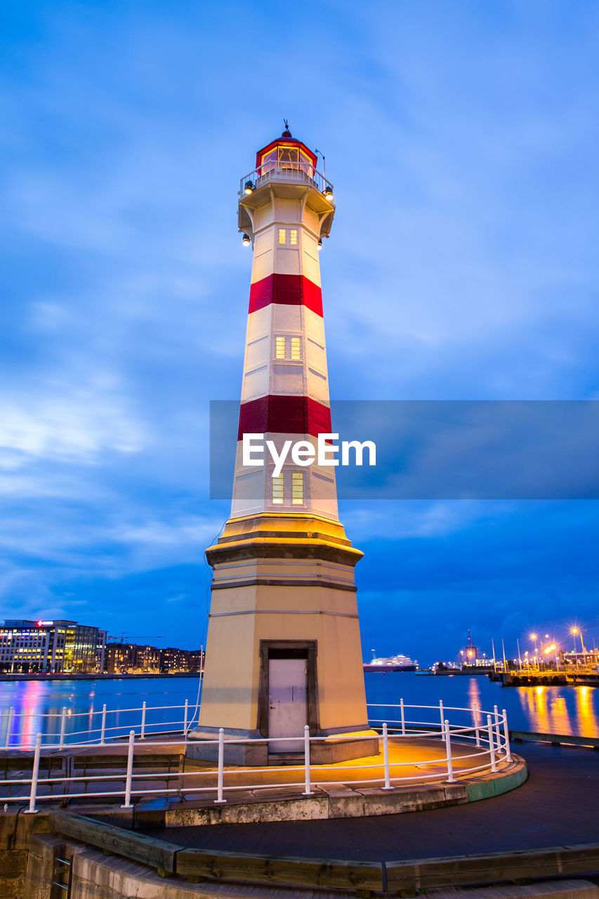 LIGHTHOUSE BY ILLUMINATED BUILDING AGAINST SKY