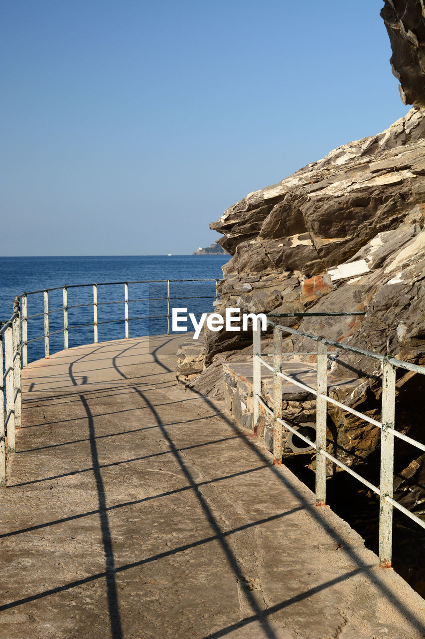 sea, water, sky, nature, coast, vacation, land, beach, clear sky, scenics - nature, horizon over water, ocean, horizon, beauty in nature, shore, tranquility, blue, sunlight, day, tranquil scene, railing, no people, sunny, travel destinations, wood, architecture, outdoors, rock, footpath, travel, walkway, holiday, pier, trip, tourism, idyllic, summer, non-urban scene, coastline, sand, seascape, built structure, shadow, the way forward, bay