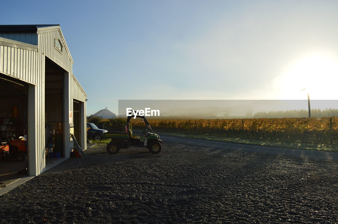 Land vehicle on road by barn against sky during sunny day