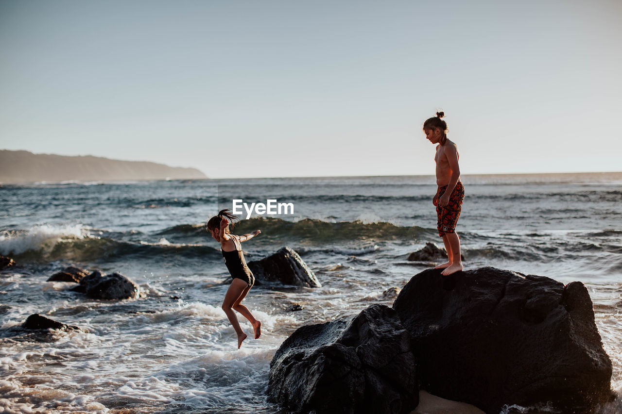 FULL LENGTH OF SHIRTLESS MAN STANDING ON ROCK AT BEACH