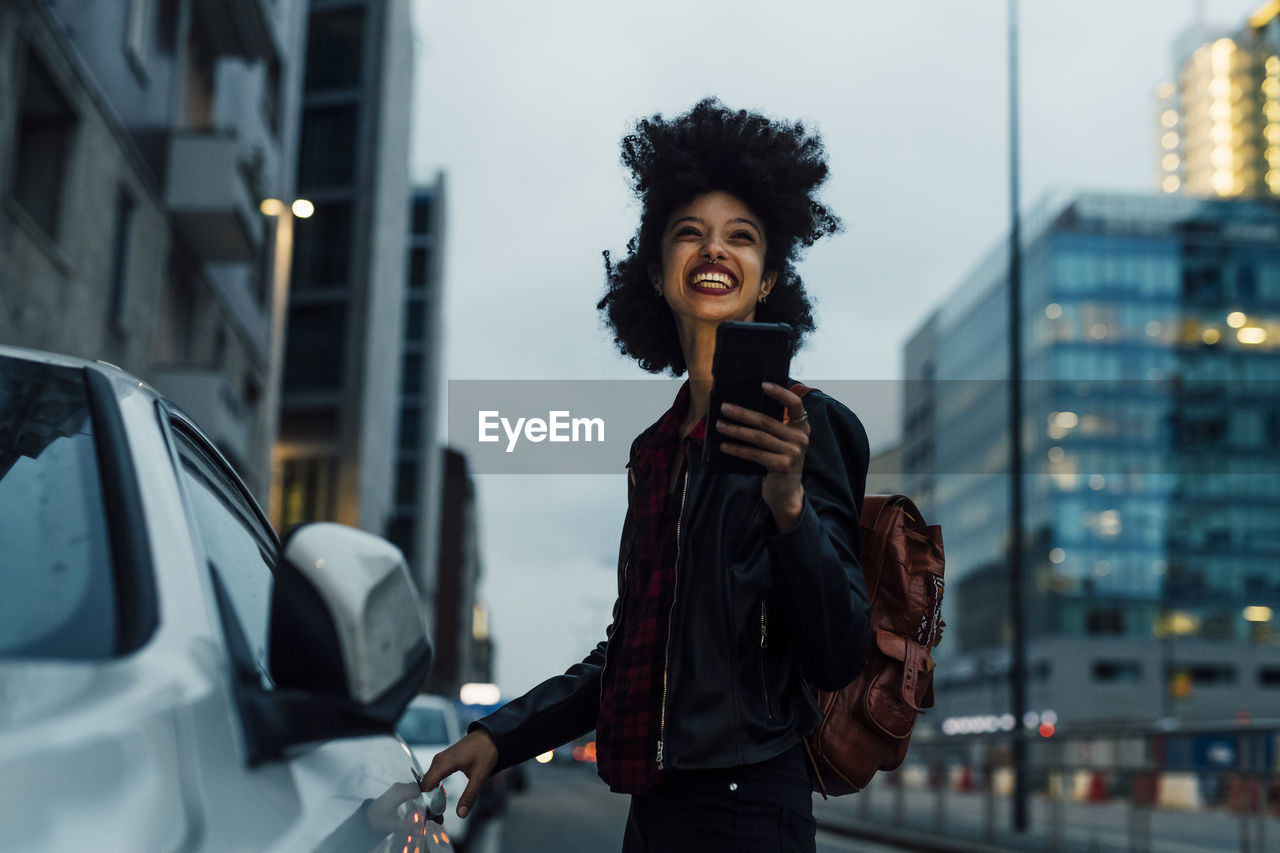 Cheerful young woman with mobile phone standing by taxi at dusk