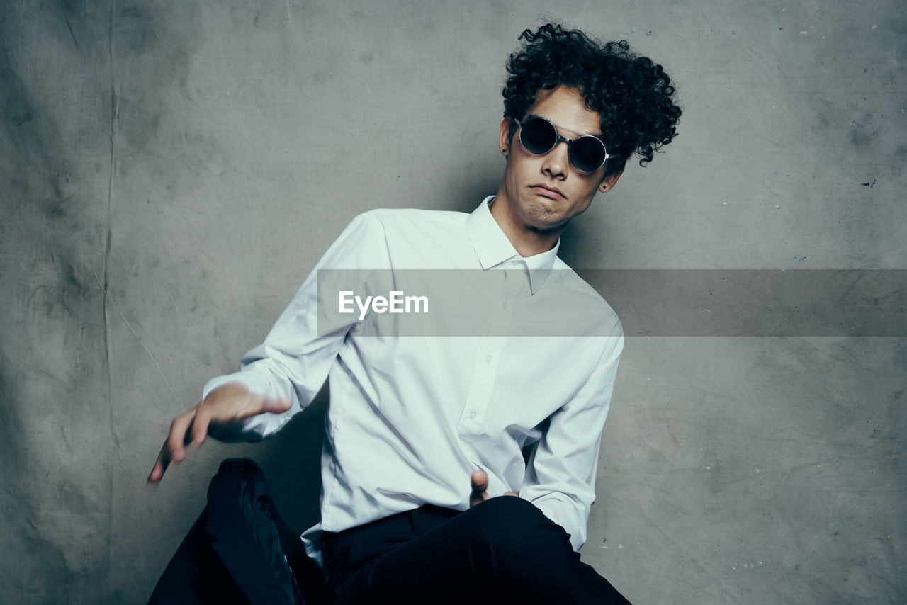 PORTRAIT OF YOUNG MAN SITTING IN SUNGLASSES AGAINST WALL