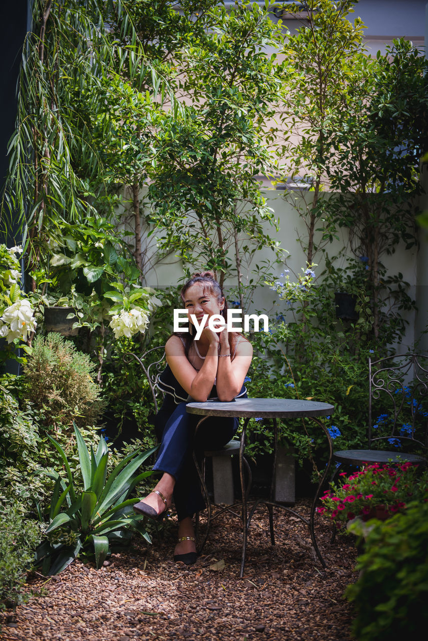 PORTRAIT OF WOMAN SITTING AGAINST PLANTS AND TREES