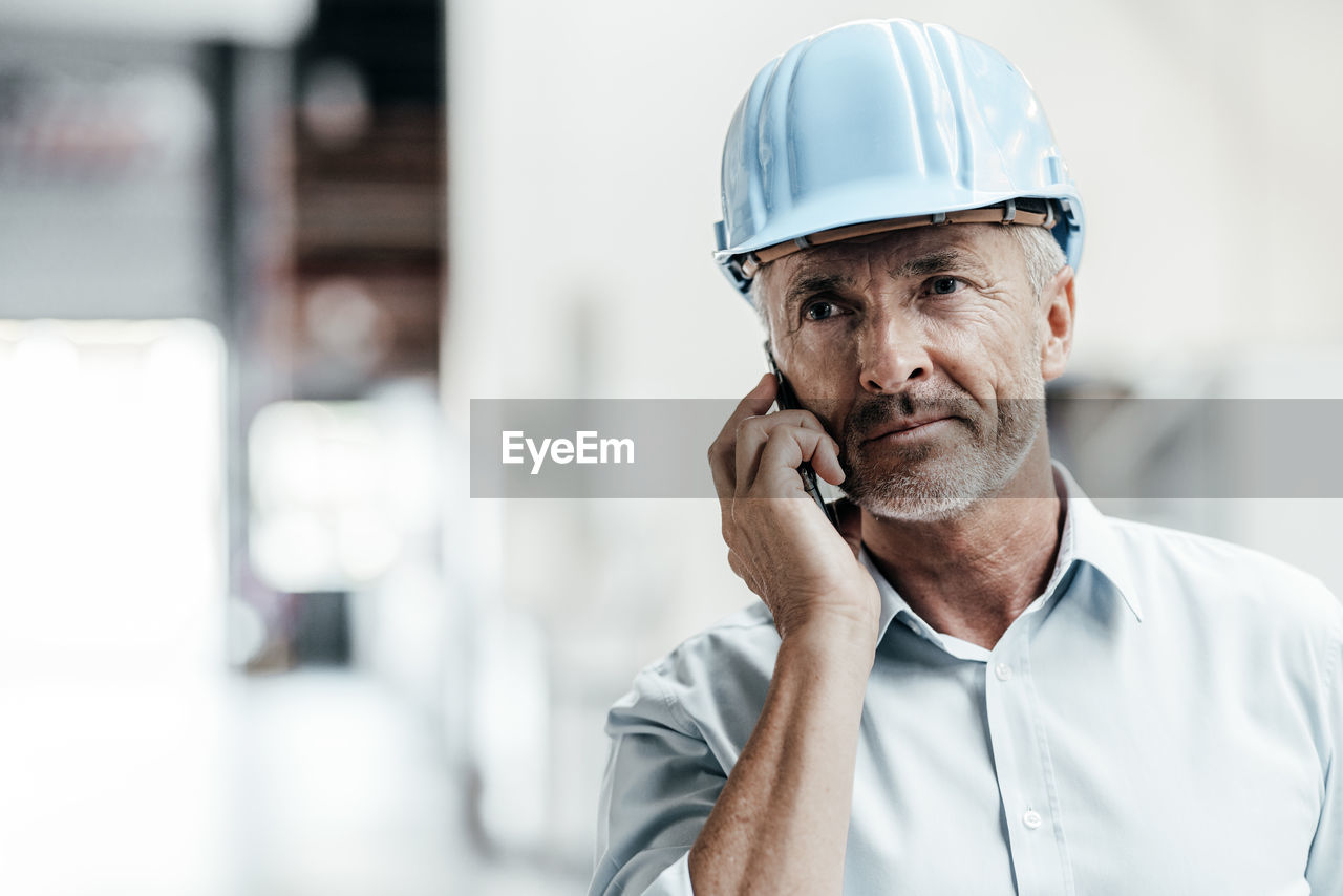 Mature male engineer in hardhat looking away while talking on mobile phone in factory