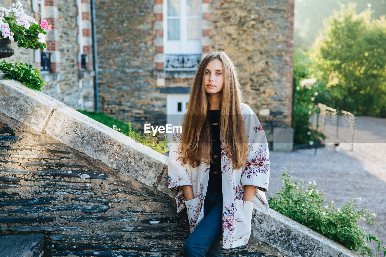 PORTRAIT OF YOUNG WOMAN STANDING AGAINST BUILT STRUCTURES