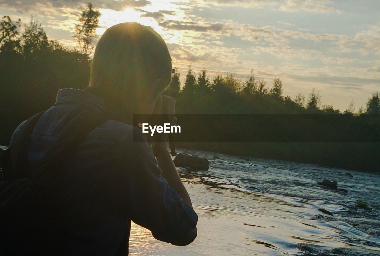 Rear view of man clicking photograph of river with camera against sky during sunset