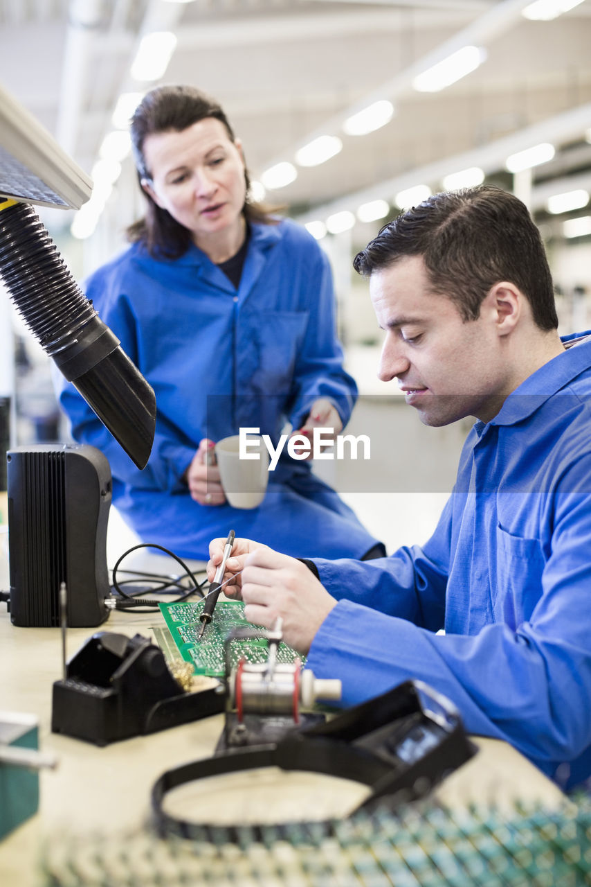 Male electrician soldering circuit board with colleague sitting on desk in industry