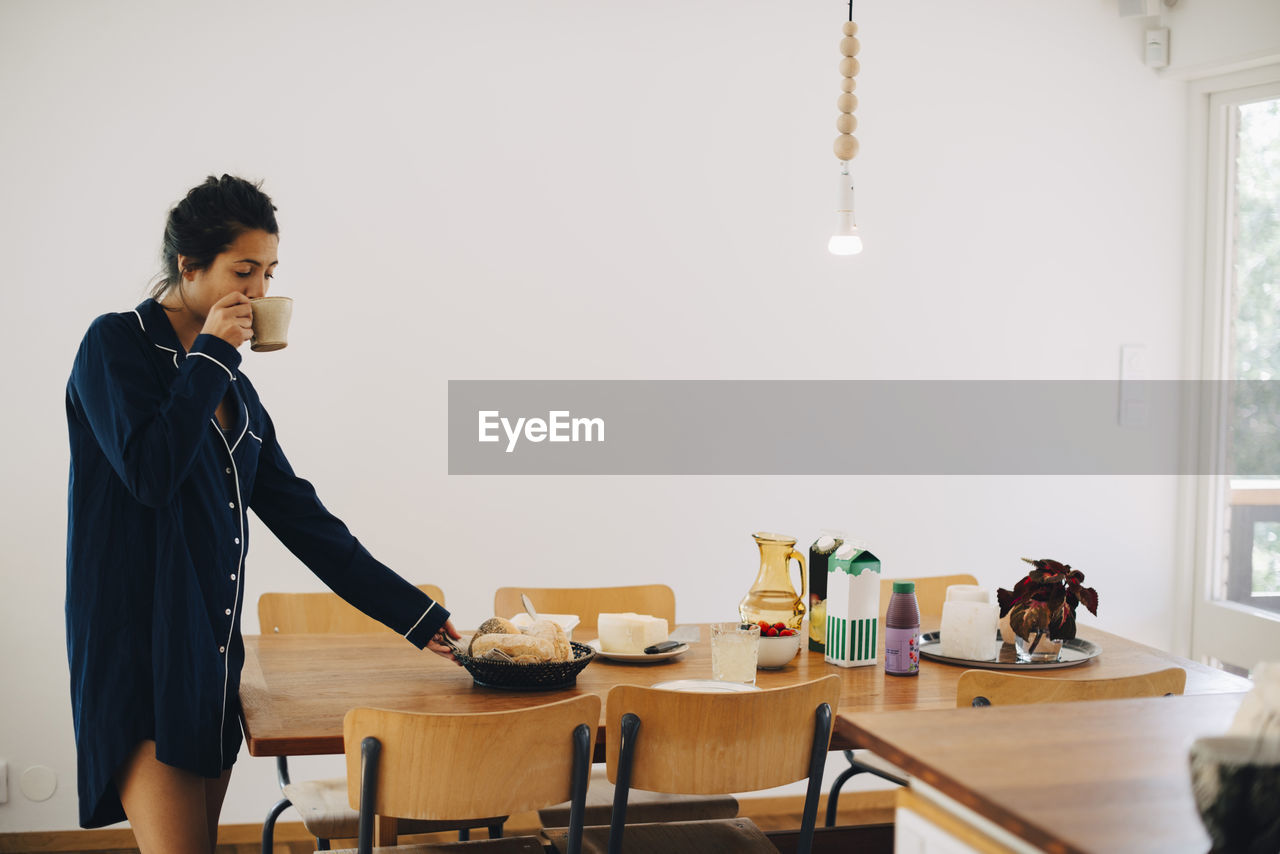 Woman drinking coffee while looking at breakfast on table by wall