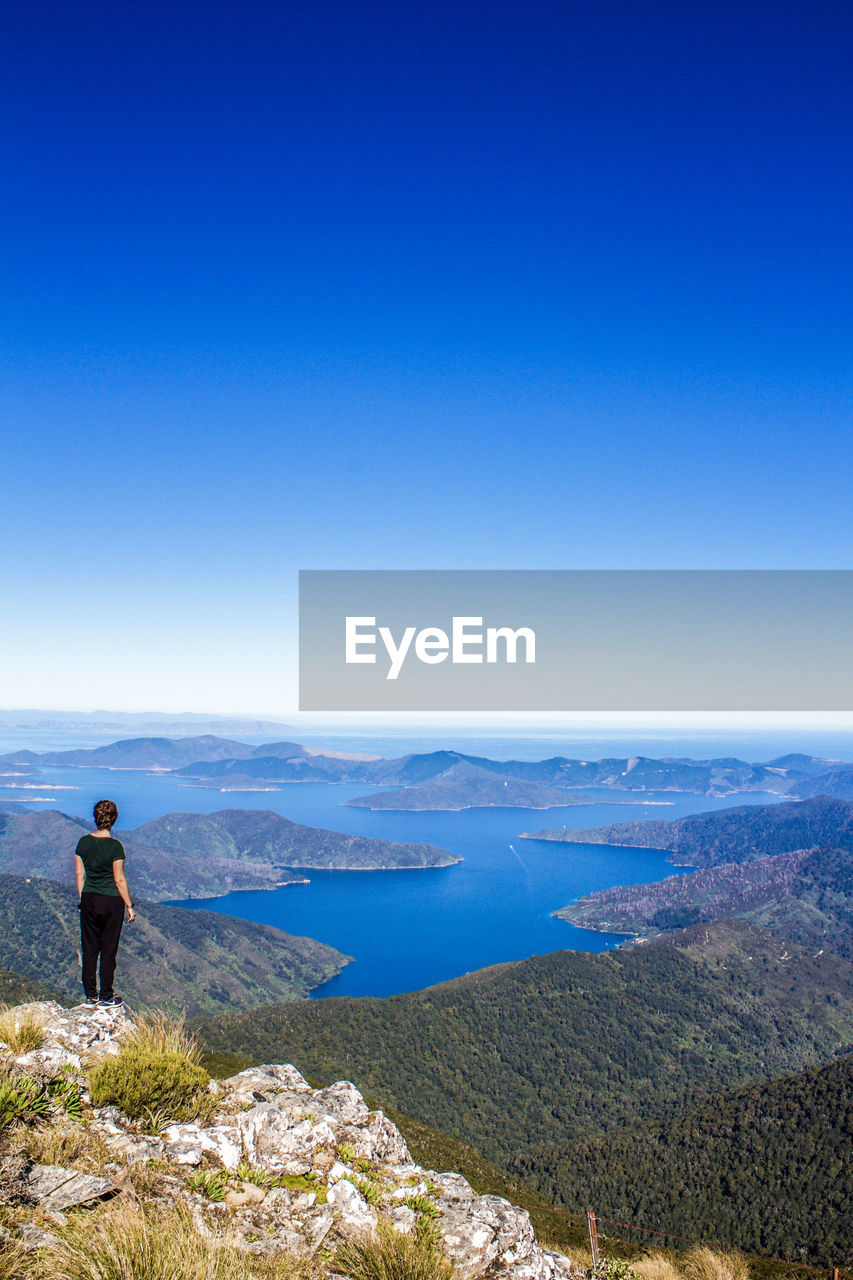 REAR VIEW OF MAN STANDING ON MOUNTAIN AGAINST SKY