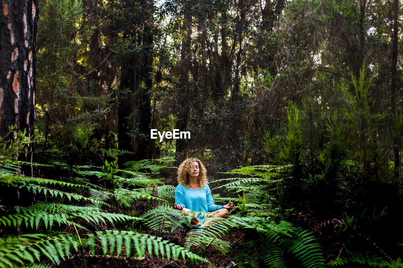 Woman with curly hair meditating while sitting amidst plants in forest