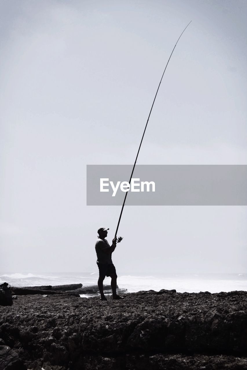 FULL LENGTH OF MAN FISHING ON ROCK AGAINST SKY