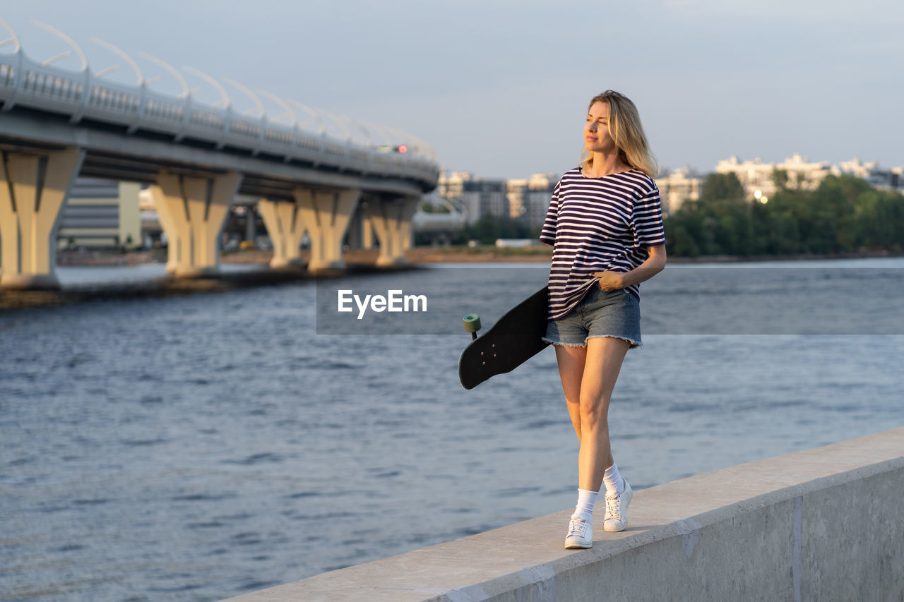 Woman walk along on river embankment hold longboard enjoy summer sunset in city. urban lifestyle