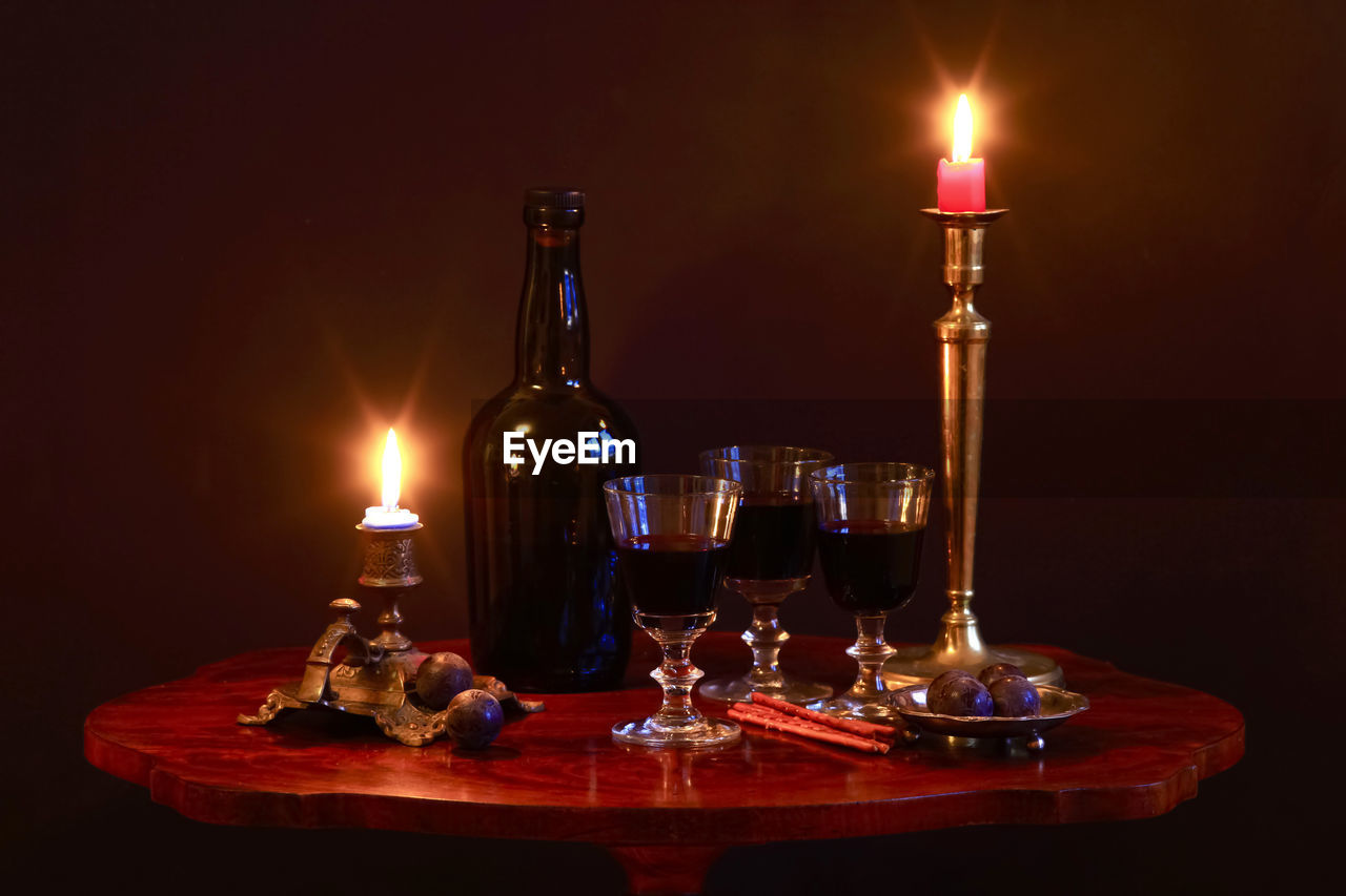 Studio shot of bottle of red wine, three filled wineglasses, plums, salty pretzels and candles burning in antique candleholders standing on small coffee table