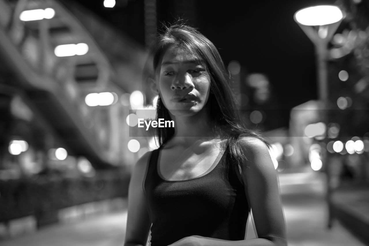 Portrait of young woman standing against illuminated city at night