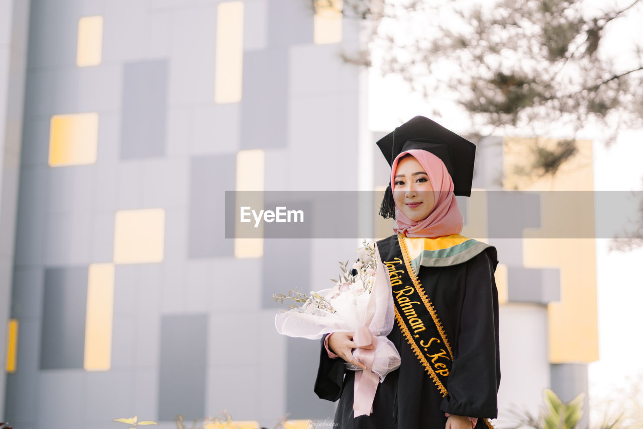 Portrait of young woman wearing graduation gown holding bouquet outdoors