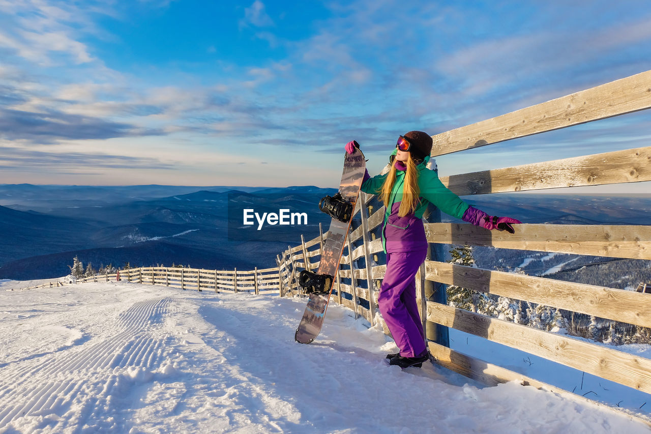 Full length if woman standing by wooden fence with snowboard against sky