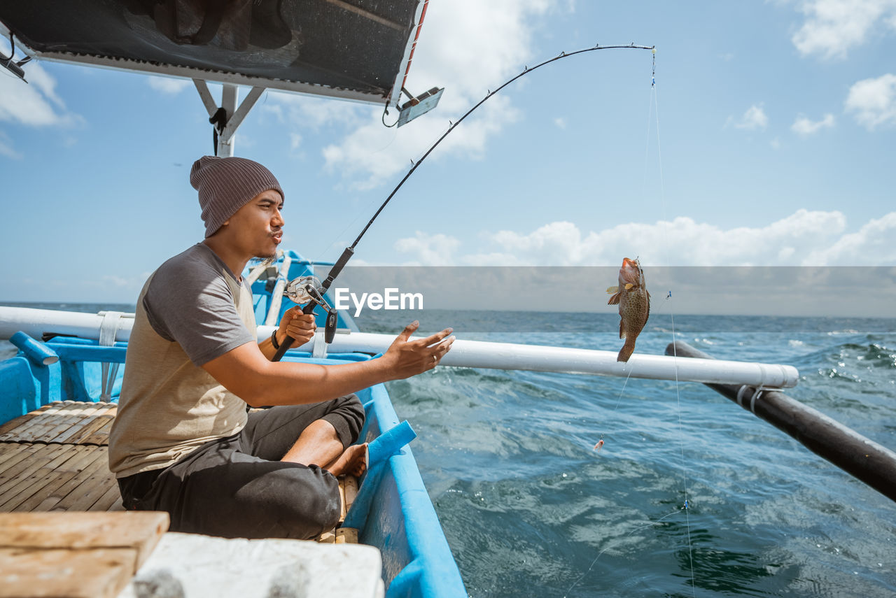 side view of man using mobile phone while sitting on boat in sea against sky