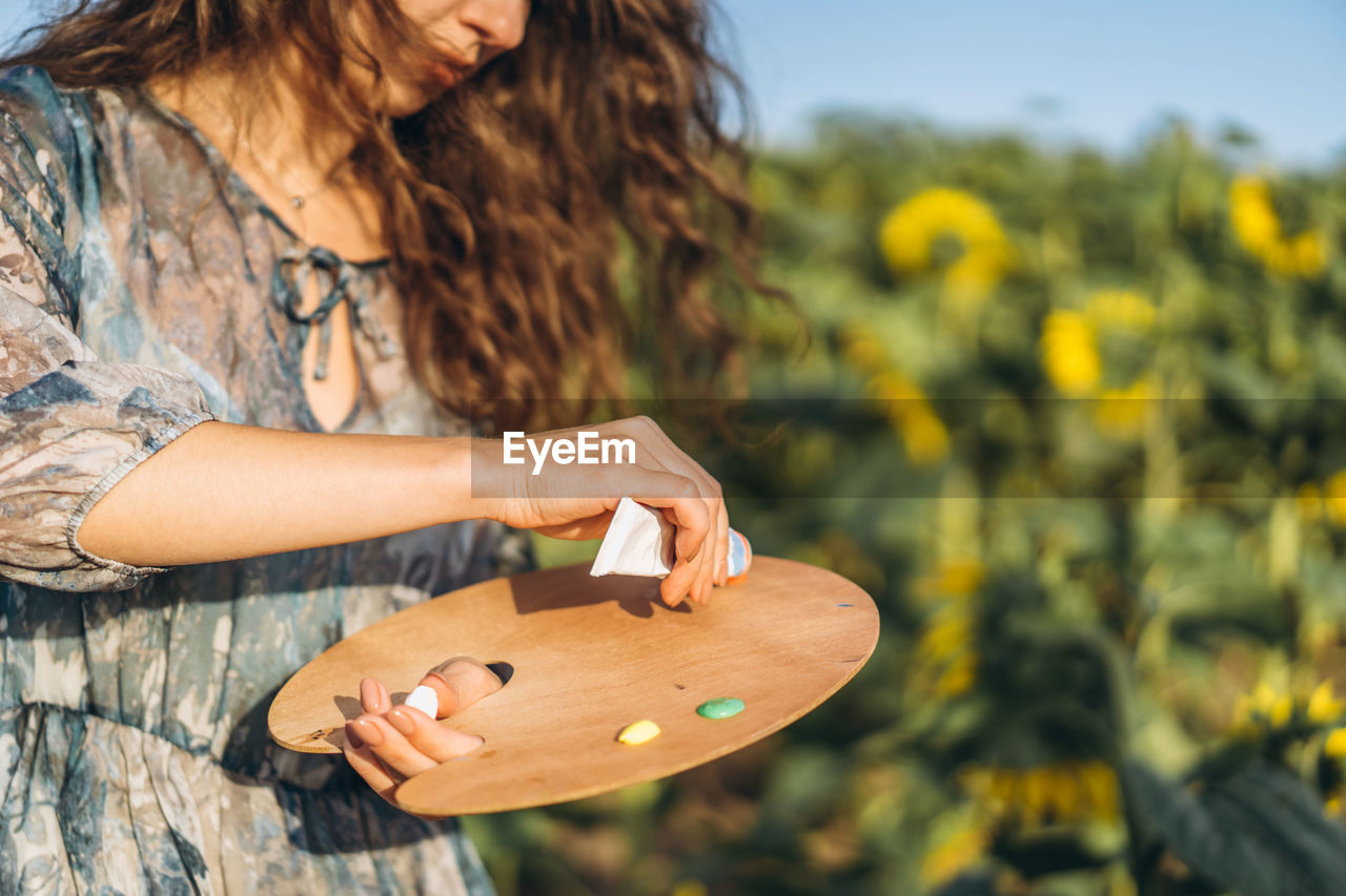 A young woman with curly hair and wearing a hat is painting in nature. 