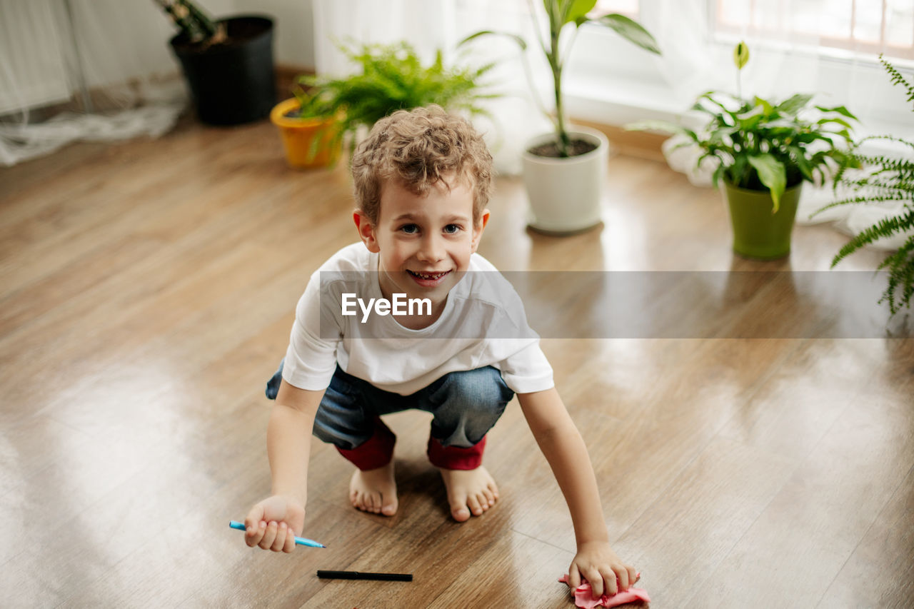 The boy is squatting on the floor in the room, holding a marker in his hand, wiping the floor