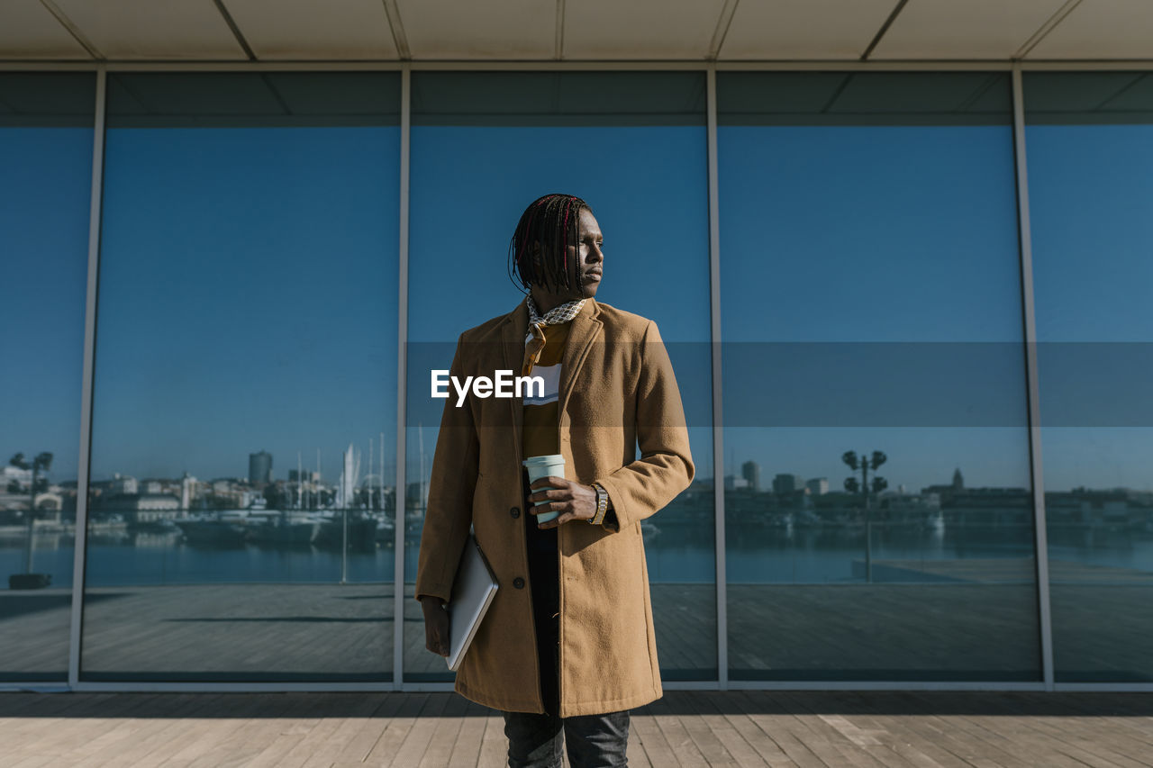 African man with laptop standing against glass wall while looking away