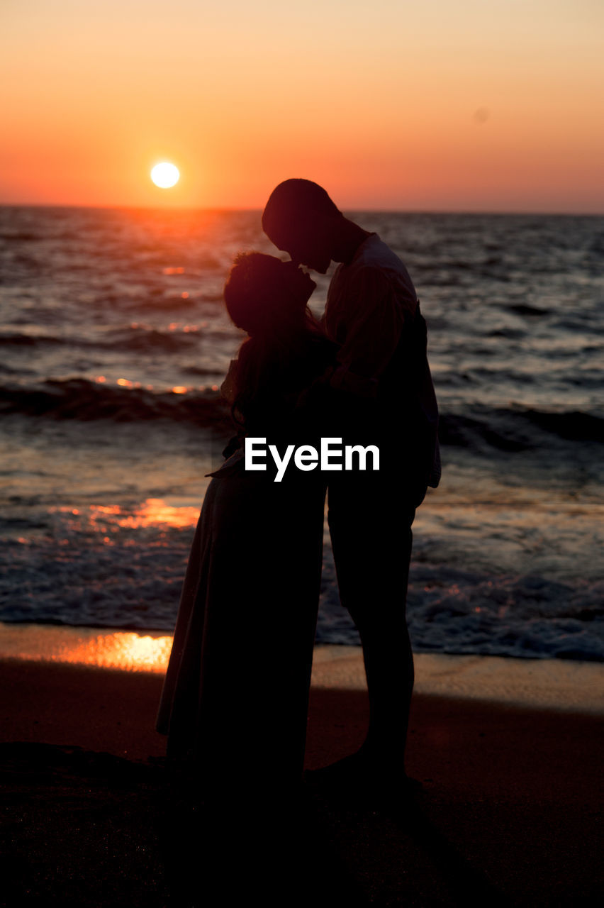 SILHOUETTE COUPLE STANDING ON BEACH AGAINST ORANGE SUNSET SKY