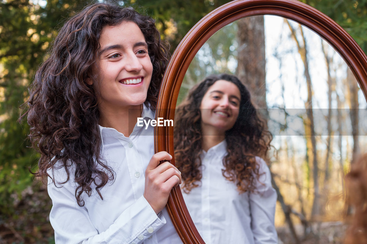 Smiling woman holding mirror with sister reflection at forest